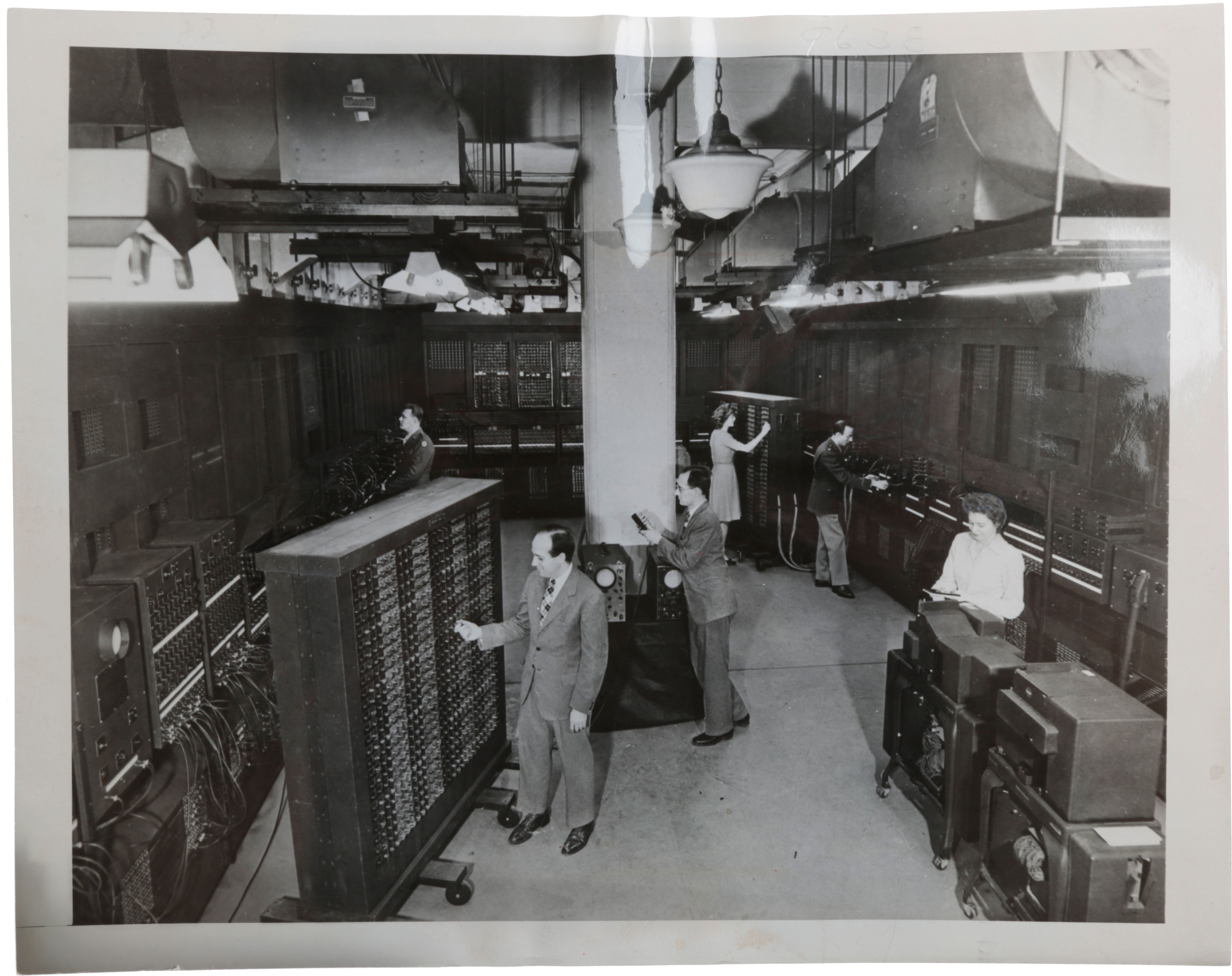 Item #5514 Photograph of ENIAC (Electronic Numerical Integrator And Computer), ca. 1945, showing Pres Eckert (centre left) and John Mauchly (centre right) working with the machine, as well as (left to right) Pfc. Homer Spence, Betty Jean Jennings, Herman H. Goldstine and Ruth Lichterman. ENIAC.